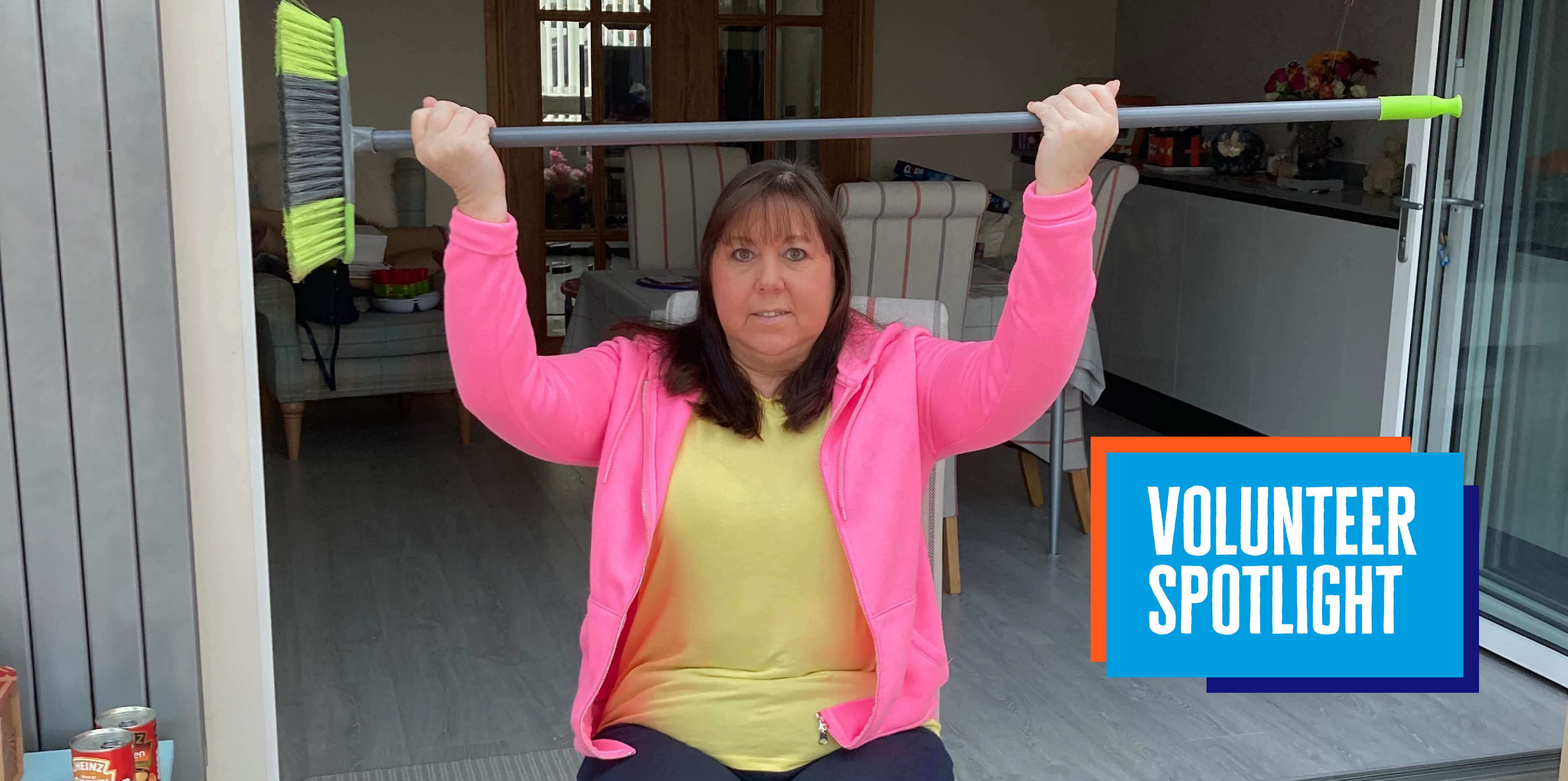 Carol wearing a pink hoodie and a yellow tshirt, sitting on a chair and lifting a broom over her head as she delivers an exercise class at home