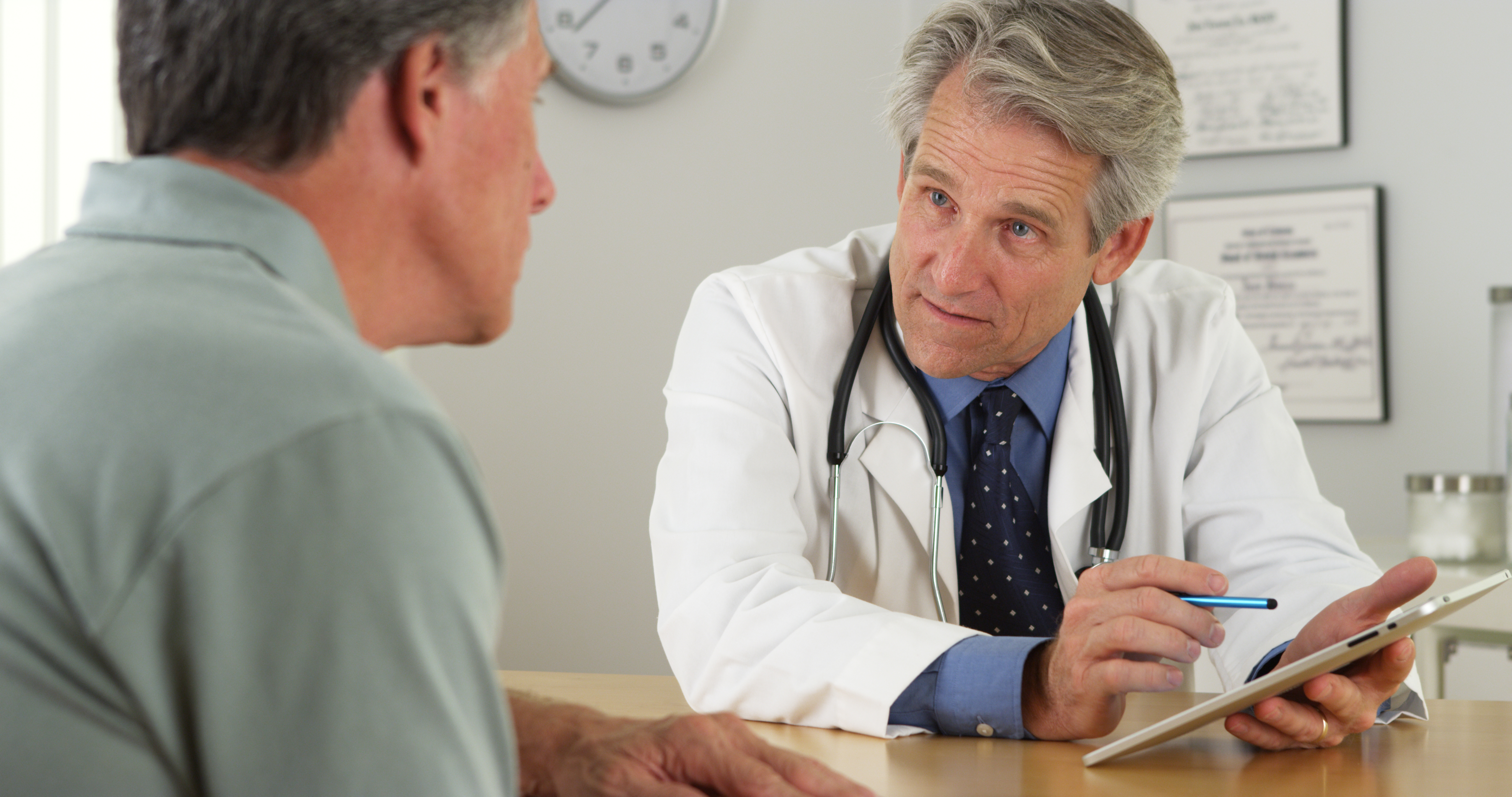 A doctor in a lab coat holds a clipboard as he talks to a male patient sat next to him.