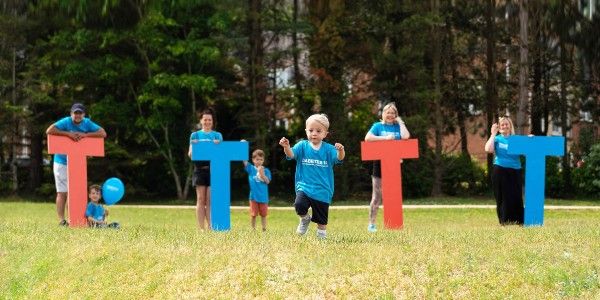 The image shows 4 giant letter Ts in a park. There is a person holding each letter and a young child running forward.