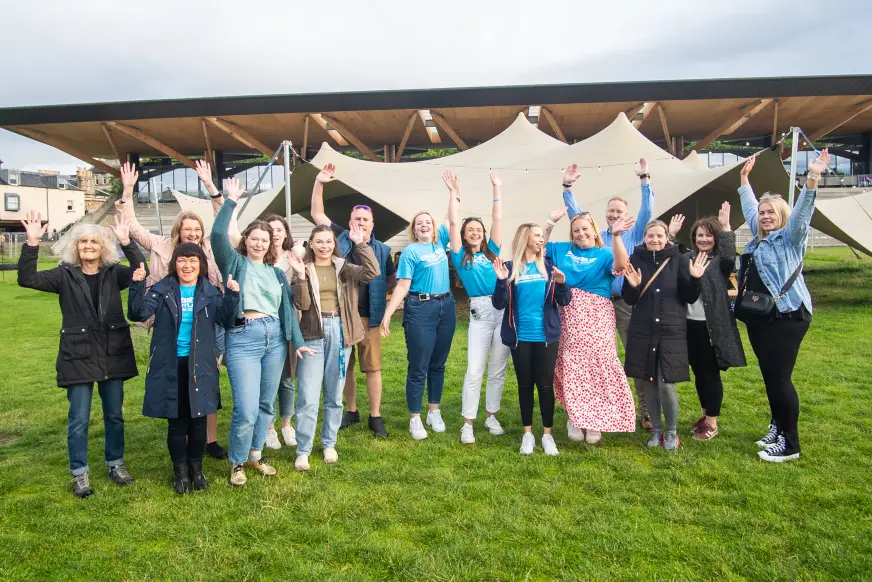 A group of people stand in a field in front of a building with their hands in the air, smiling at the camera. Some people are wearing blue Diabetes UK t-shirts.