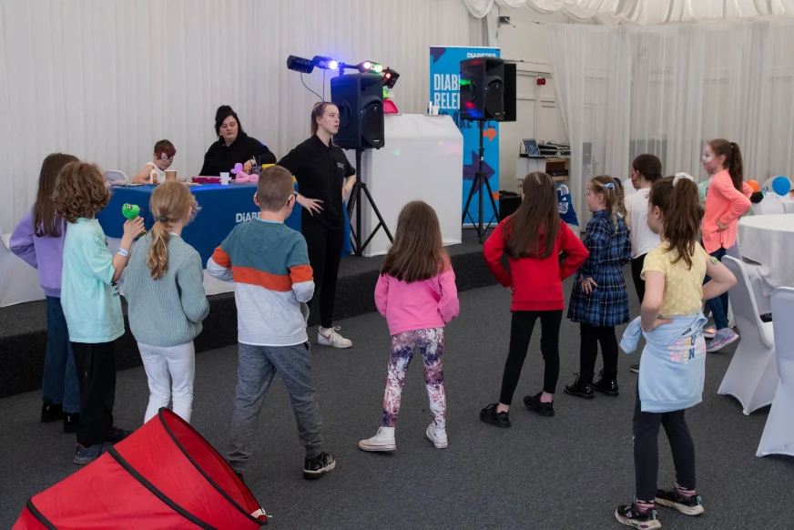 A group of children stand in a semi-circle around a woman. They all have their hands on their hips as if they are dancing. They stand in front of a Diabetes UK desk and banner.