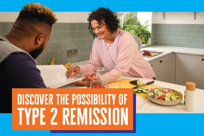 A smiling woman leans across a breakfast bar with fresh vegetable around her. She is reading a recipe book. A man sits across from her.