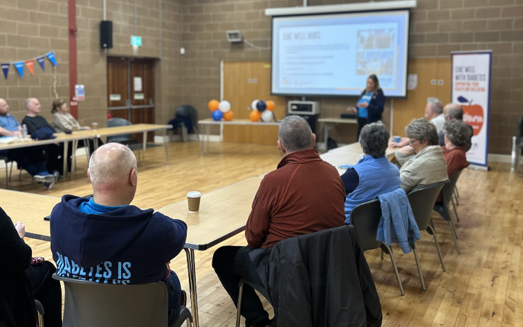 A group of people sit around at a long table listening to a Diabetes UK member give a presentation. 