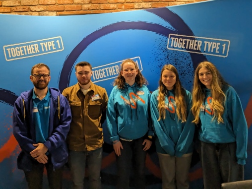 A group of 5 people stand in a line against a Together Type 1 wall smiling at the camera. There are Diabetes UK staff members at either end wearing blue Together Type 1 hoodies.