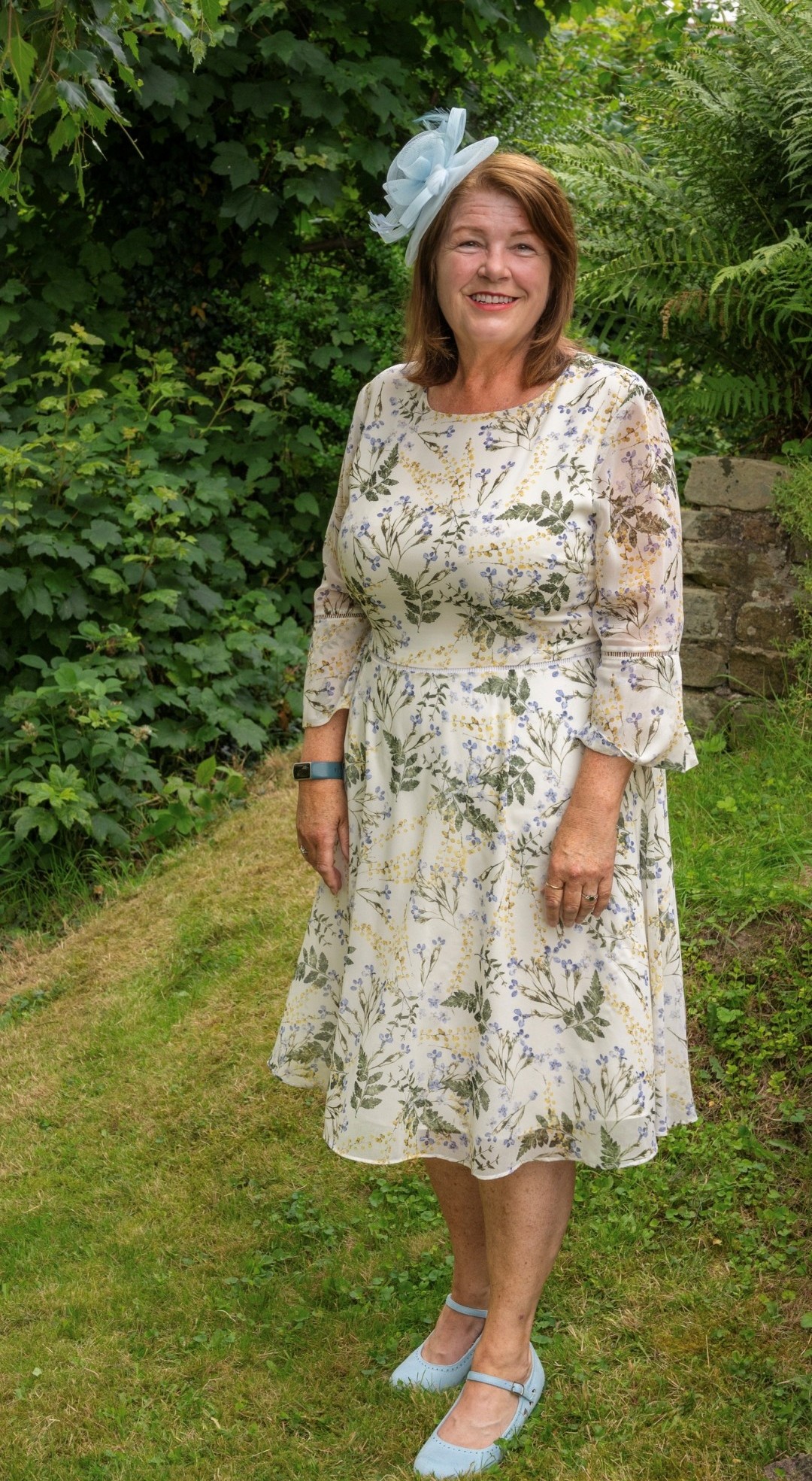 Denise, who has shoulder length auburn hair, pictured outdoors on a family members wedding day. She is wearing a floral dress, blue head piece and blue shoes.