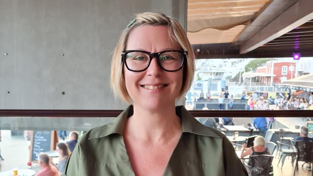 A photo of Nicola smiling to camera, wearing black glasses, with a cafe in the background