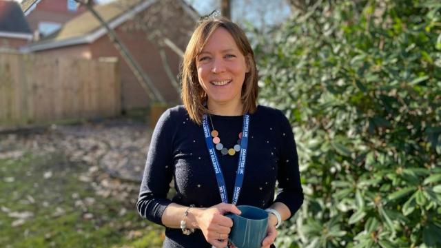 A photo of Jenny standing in her garden, smiling to camera and wearing a Diabetes UK lanyard while holding a mug