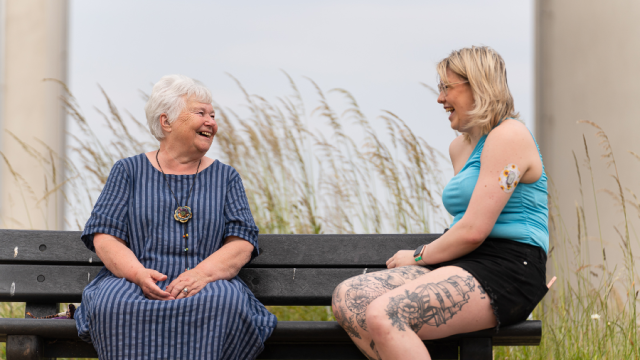 The image shows a lady in a dark blue dress with grey hair sitting on a bench next to a lady in a blue t-shirt and black shorts with blonde hair