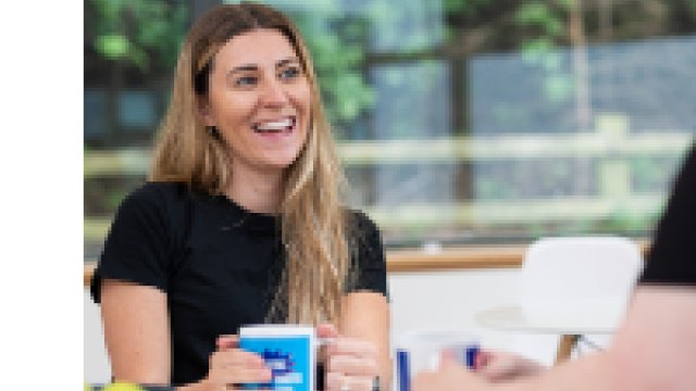 The image shows a lady with long hair laughing with her hands wrapped around a mug. 