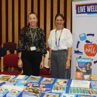 The image shows two ladies next to a banner which reads 'Live Well Hub'. They are standing behind a table with leaflets about diabetes and living well. 