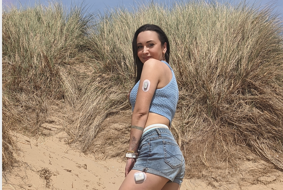 Natalie stands on a beach, with diabetes tech on her arm and leg