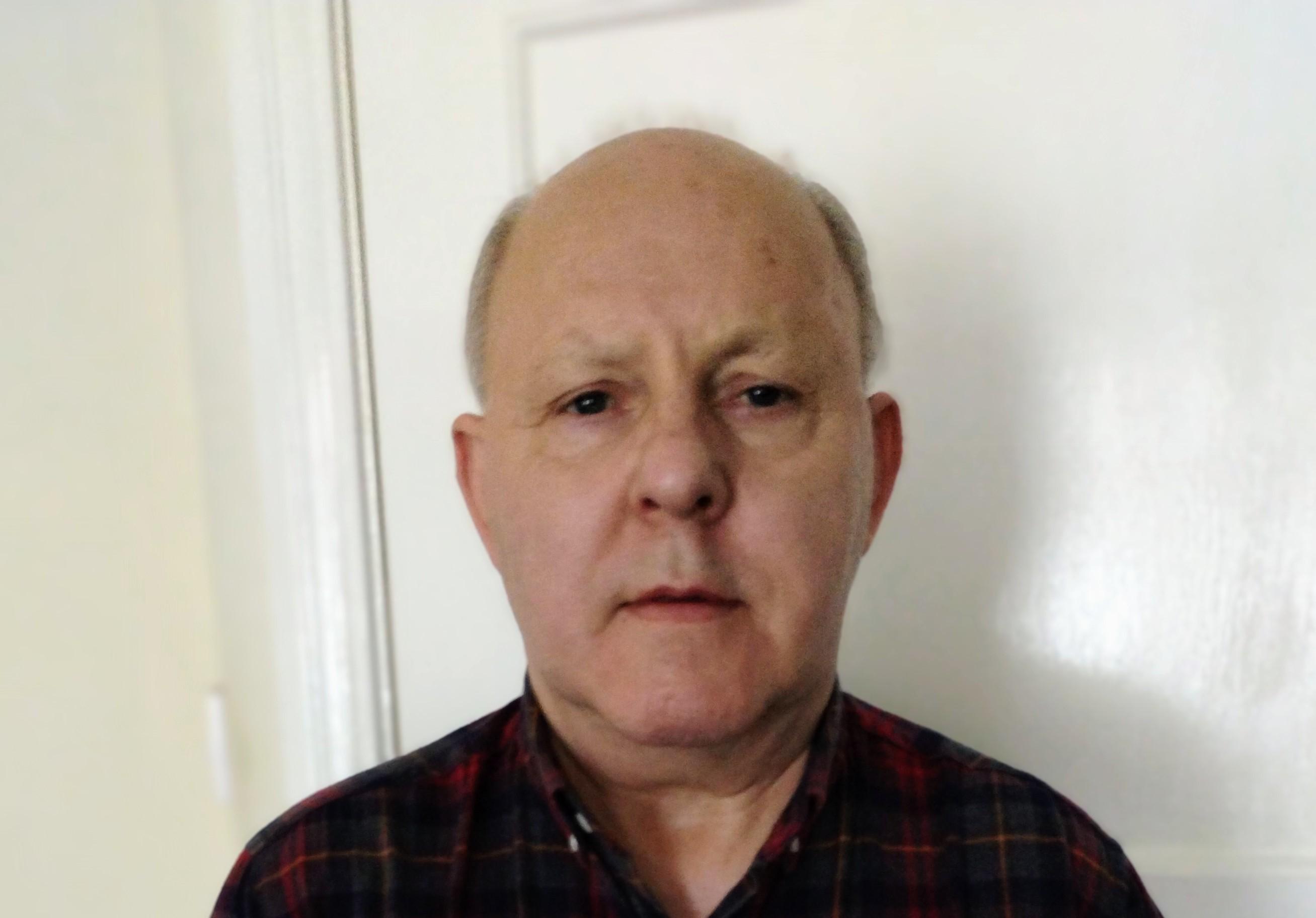 A man wearing a checked shirt, standing by a wall