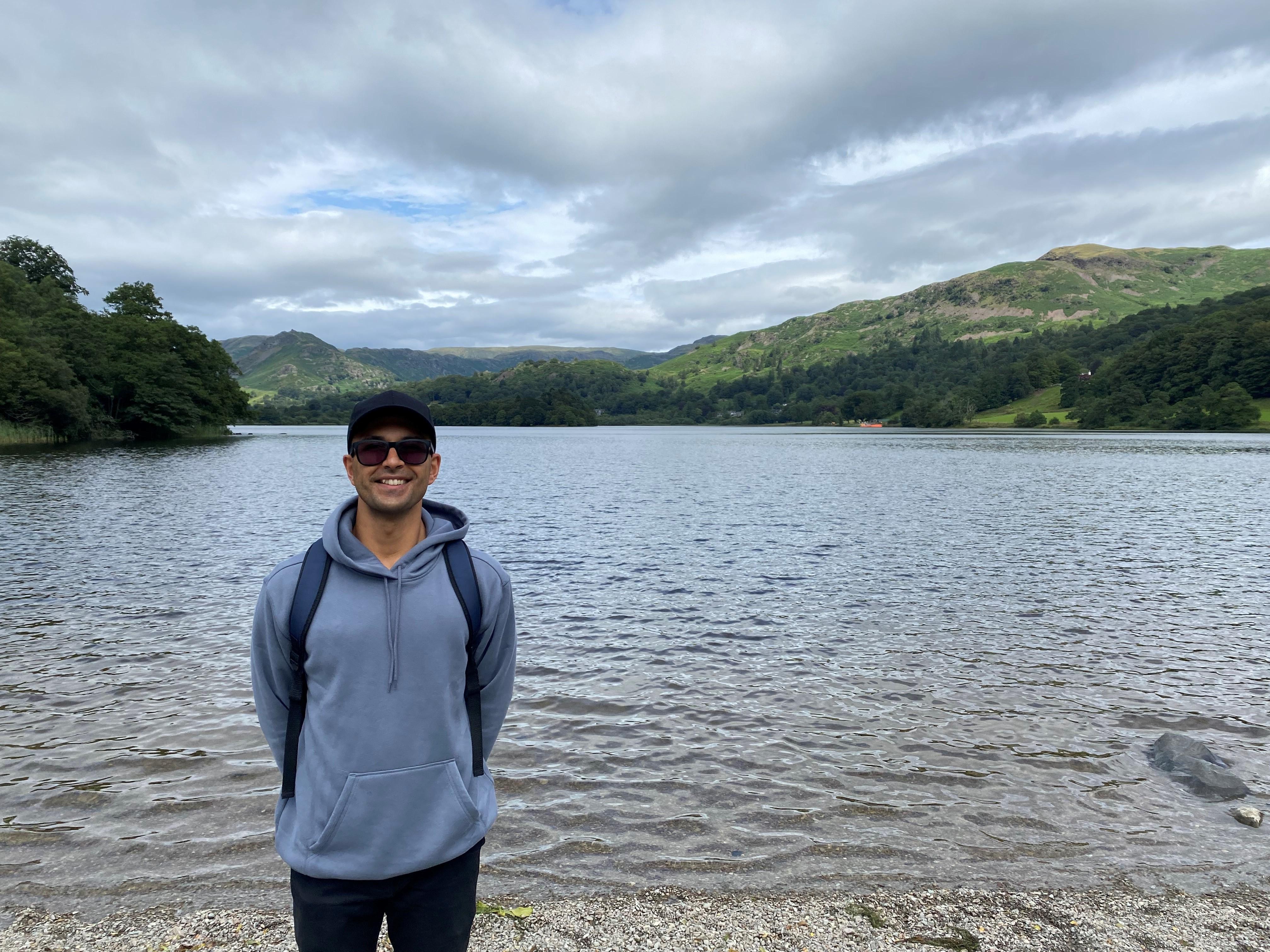 A man wearing sunglasses stands by a lake, smiling