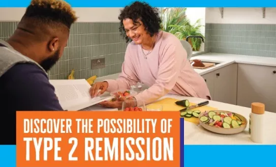 A smiling woman leans across a breakfast bar with fresh vegetable around her. She is reading a recipe book. A man sits across from her.