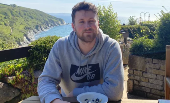 Rob sits at a table outdoors with a bowl of food in front of him. He's looking at the camera with the sea and hills behind him.