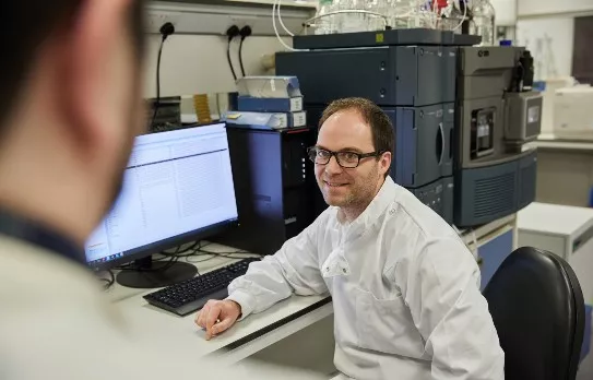 a man in a lab coat sitting at a desk next to a computer with lines of text on it, talking to a colleague off-camera