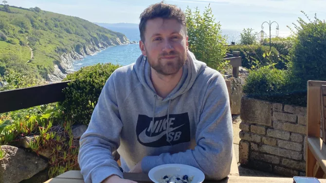 Rob sits at a table outdoors with a bowl of food in front of him. He's looking at the camera with the sea and hills behind him.