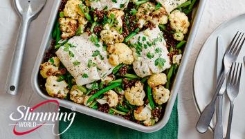 Fish traybake with cauliflower and lentils and parsley sprinkled over the top. There is a serving spoon and a white plate with two forks and a butter knife on the side.