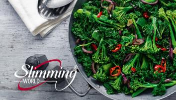 A metal skillet against a grey table filled with long-stemmed broccoli, kale, green peppers, red chillies and red onion