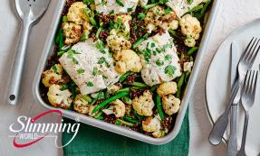 Fish traybake with cauliflower and lentils and parsley sprinkled over the top. There is a serving spoon and a white plate with two forks and a butter knife on the side.