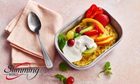 A small baking tin with oats, mixed fruit, yogurt, and mint leaves. A cloth napkin with a spoon on top is to the left of the baked oats.