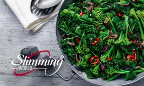 A metal skillet against a grey table filled with long-stemmed broccoli, kale, green peppers, red chillies and red onion