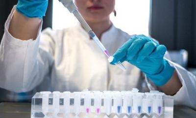 A woman in a white lab coat and blue gloves using a pipette to put liquid in to to test tubes