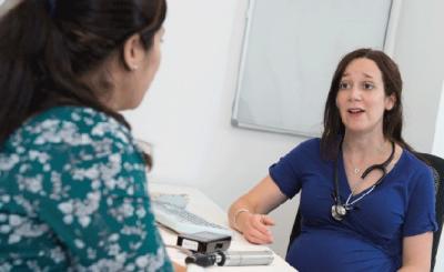 A GP wearing a blue dress, with a stethoscope around her neck sitting at a desk, facing the camera, talking to a patient with her back to camera